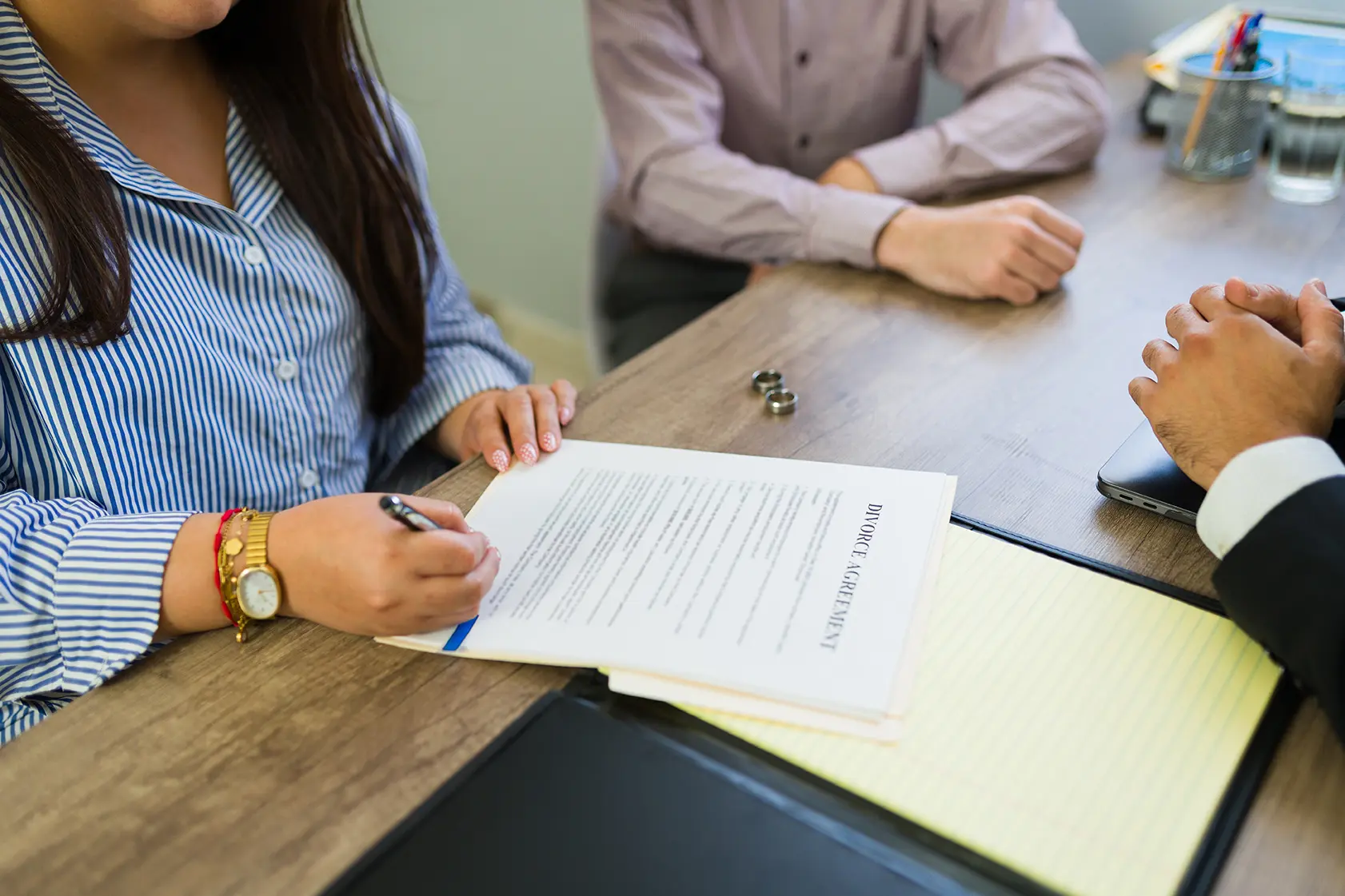 Closeup of woman signing legal papers with a lawyer present, preparing to end a marriage. Two wedding rings lay on the table